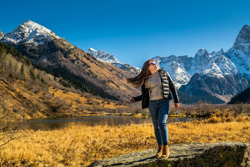 Naklejka na ściany i meble Beautiful young russian woman in front of autumn landscape with mountain lake Tumanly-kel, the Mist lake, located in Russia, near Dombay , in Caucasus mountains, Gonachkhir gorge