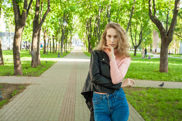 blonde girl with curly hair in blue jeans and a black leather jacket, standing in the Park on the background of the path and trees with a place for the inscription