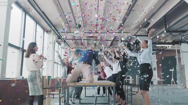 Manager start to dance on the table, sings into a megaphone, accelerates and slide on lap. Colleagues blow up flappers with confetti. Employees celebrate success. Corporate party business team