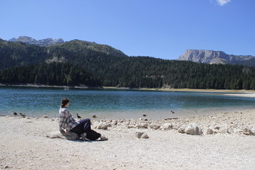 Fototapeta na wymiar Paradise views of the national park Durmitor in Montenegro. Turquoise water of the lake, pine forest and mountains. Stunning background with nature girl tourist sitting on the beach