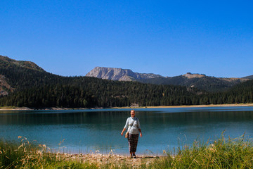 Fototapeta na wymiar Paradise views of the national park Durmitor in Montenegro. Turquoise water of the lake, pine forest and mountains. Stunning background with nature tourist girl rejoicing on the beach