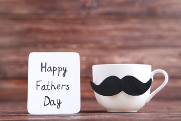 Text Happy Fathers Day and cup with black mustache on wooden table