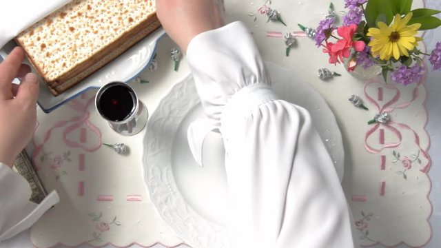Female hands had just pulled the matzo out of the pack and put it on a white plate. The table is covered with a holiday tablecloth. Glass of red wine is on the left, flowers are in a vase on the right