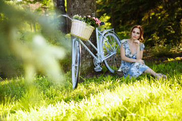 portrait of a beautiful girl in the forest, sitting on the grass, next to the bike, with a basket of flowers, behind the rays of the sun, a blue flowered dress, summer walk.
