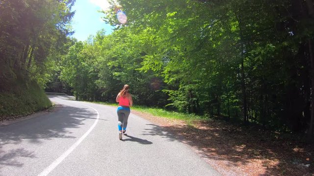 Wide forward tracking shot of a woman with a long pony tail running on the sunny road through forest