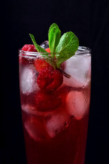 Red drink with ice, raspberries and mint in a tall glass against the black background