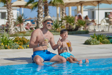 Fototapeta na wymiar Dad and son enjoying their summer holidays. They are holding their cocktails in the hands, while sitting near the hotel’s swimming pool.