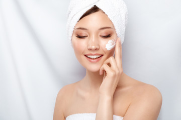 pretty girl with bath towel smilling and putting on facial cream on cheek, happy face care, body treatment, beauty concept