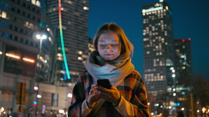 Caucasian Young Woman is Using Smartphone App Outdoors in the Downtown City in the Late Evening or Night with Beautiful Blurred Cityscape Lights of Skyscraper Windows