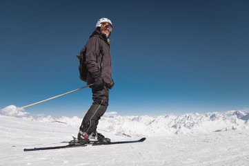 Fototapeta na wymiar A bearded male skier in a helmet and a ski mask is standing on skis against the background of snow-capped mountains and a blue sky. Athlete in a black suit