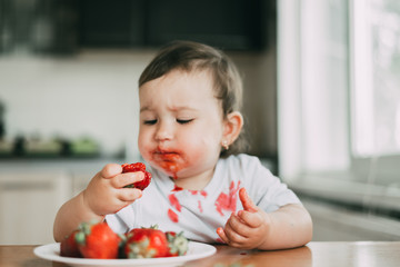 Little girl child in white t-shirt eating strawberries all smeared and dirty