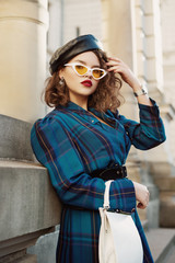 Fashionable curly woman wearing trendy leather beret, cat eye sunglasses, checkered blue dress,...