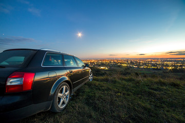Black car parked at night in green meadows on copy space background of lights of distant city buildings and bright blue sky with first star at sunset.