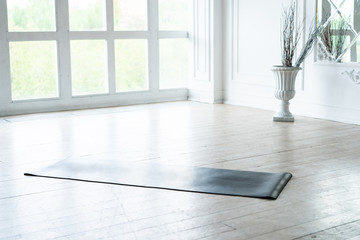 Unrolled yoga mat lying in empty light fitness studio on wooden floor, unfolded sport equipment prepared for training, modern pilates loft room ready for workout session. Healthy life concept.