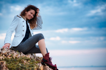 fashionable girl dressed in white jacket and wide trousers posing at sunset sky background, sits on the stone