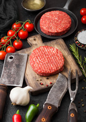 Fresh raw minced homemade farmers grill beef burgers on round chopping board and frying pan with spices and herbs and meat hatchet on black board. Oil, garlic and tomatoes.