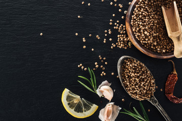 top view of coriander seeds in silver spoon and wooden bowl on black textured background with dried...