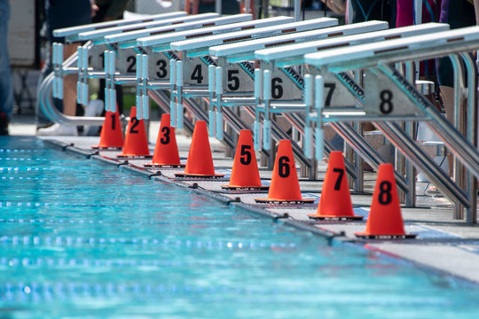 Starting blocks at the edge of a competition swimming pool.  Sunny afternoon.  No swimmers visible.  Orange cones with numbers 1-8.  Numbers on the blocks.
