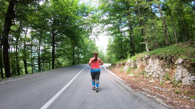 Forward tracking shot of an overweight woman running on an asphalt road in the park