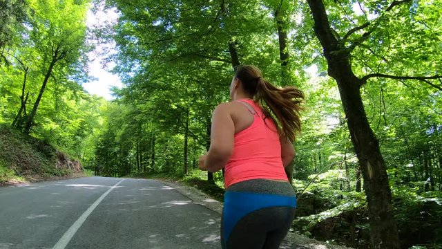 Medium forward tracking shot of an overweight woman running in sunshine on a road through a forest