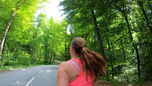 Medium tracking shot of an overweight woman with a pony tail running in the sunny park