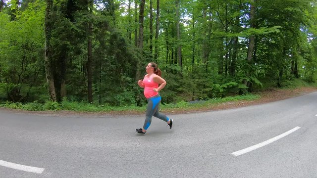 Tracking shot of an overweight woman running on the asphalt road surrounded by lush forest