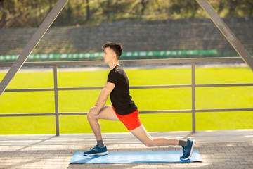 Fototapeta premium Young man training yoga outdoors. Sporty guy makes stretching exercise on a blue yoga mat, on the sports ground.