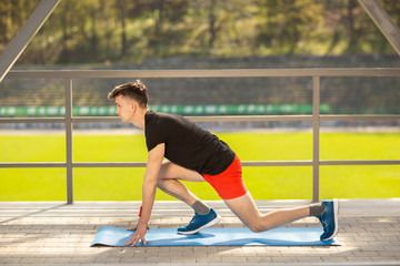 Fototapeta premium Young man training yoga outdoors. Sporty guy makes stretching exercise on a blue yoga mat, on the sports ground.