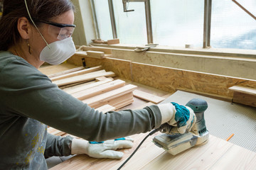 Young woman grinds the wood board with angular grinding machine at workshop