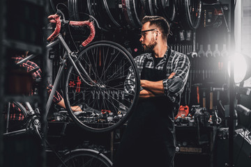 Fototapeta na wymiar Handsome bearded man in glasses is standing near fixed bicycle at his own workshop.