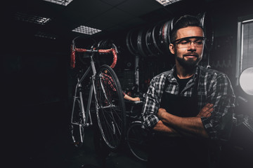 Fototapeta na wymiar Handsome bearded man in glasses is standing near fixed bicycle at his own workshop.