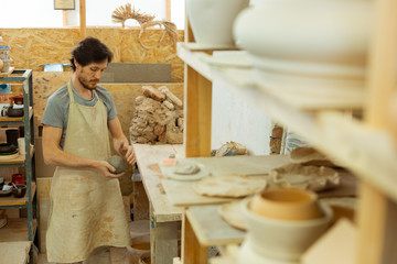 Concentrated dark-haired guy in long apron rolling clay