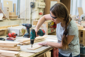 Young woman with drill making hole into wooden plank at workshop