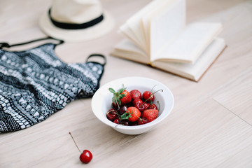 Top view travel or vacation concept. Swimsuit, hat, book and fresh sweet cherries and strawberries on wooden background.  Flatlay. Summer background. 