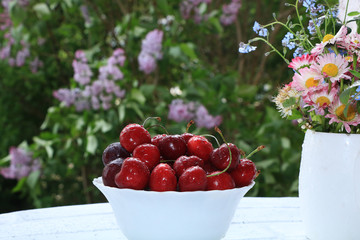 A cup of cherries on a sunny table in a flowered garden, selective focus. Sweet cherry with droplets and a branch of lilac in the background in the garden,