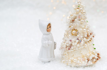 A little doll in winter clothes is looking at the New Year tree. Christmas tree with pearls and beads beautiful bokeh background.