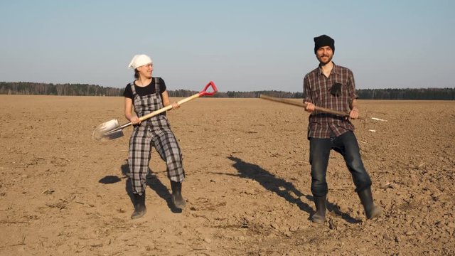 Two funny farmers man and woman with shovel and pitchfork dances on freshly plowed field. Happy farming concept.