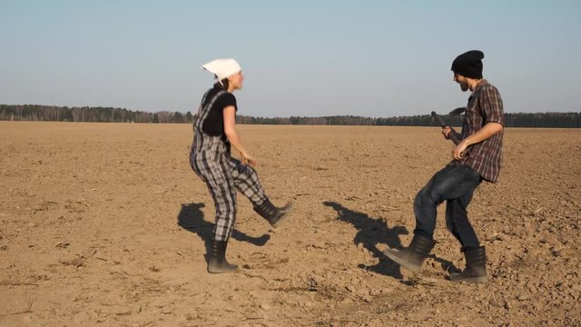Two funny farmers man and woman holds shovel and pitchfork like a guitar and dances on freshly plowed field. Happy farming concept.