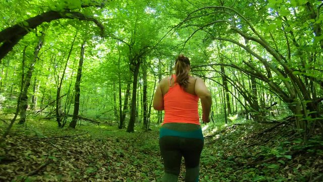 Forward tracking low angle shot of an overweight woman running up a forest path with sun shining through the trees