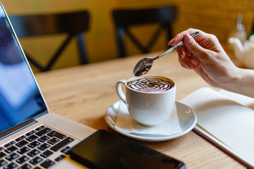 Women's hands holding a spoon over a Cup of coffee with pattern on the foam. Hot freshly brewed coffee in a white mug next to the smartphone and laptop on a wooden table in the interior of the cafe.