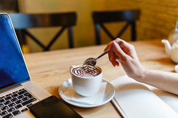 Women's hands holding a spoon over a Cup of coffee with pattern on the foam. Hot freshly brewed coffee in a white mug next to the smartphone and laptop on a wooden table in the interior of the cafe.