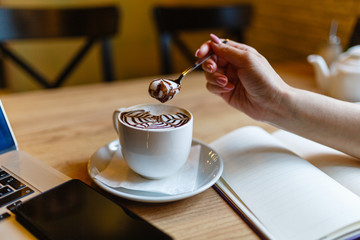 Women's hands holding spoon with foam over Cup of coffee with a pattern on the foam. Girl eats the foam from the cappuccino. Hot coffee in white mug next to the smartphone and laptop on wooden table.