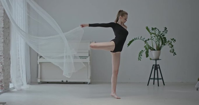 Young blond woman performing graceful dance moves while holding the filmy fabric of a window drape in a bright white studio. RAW video record.