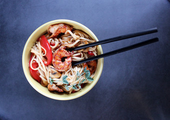 udon noodles with shrimps and vegetables in a yellow bowl with chopsticks on a dark background