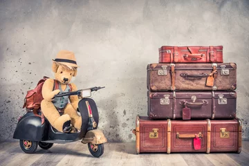 Selbstklebende Fototapeten Retro Teddy Bear toy in tourist hat sitting on old rusty children's pedal scooter from 60s, leather backpack and outdated trunks luggage, antique valises. Travel concept. Vintage style filtered photo © BrAt82