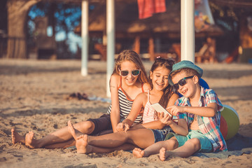 Portrait of positive children laughing watching funny video on smartphone on sandy beach