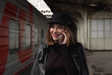 Obraz premium Attractive young woman millenial in black clothes and a hat at the railway station next to the train. Speaks on a mobile phone, smiles and waits for the train.