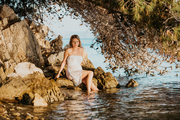 Beautiful young pregnant woman with white wet dress relax on the rocks near the sea water. Summer at sunset. Concept Love and pregnancy