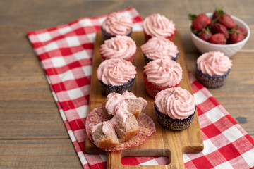 Homemade Strawberry Cupcakes on Wooden Cutting Board with one Open; Bowl of Fresh Strawberries Nearby; Red and White Checked Napkin on Wooden Table