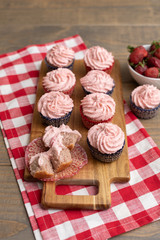 Homemade Strawberry Cupcakes on Wooden Cutting Board with one Open; Bowl of Fresh Strawberries Nearby; Red and White Checked Napkin on Wooden Table